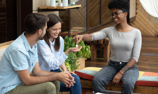 Happy couple sitting on a couch holding out their hands to receive keys to a home